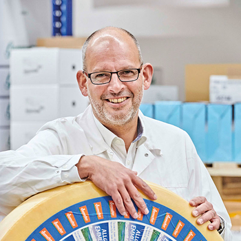 The image features a smiling man in a white lab coat leaning against a large wheel covered in stickers.